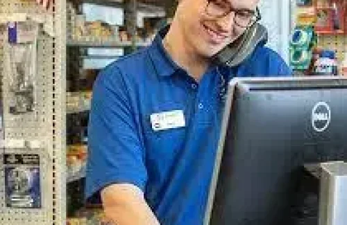A Man in an auto store, standing behind a desk, on the phone, smiling, looking at a computer screen.